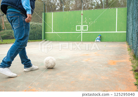 A quiet scene on a fenced playground. A child in blue sits safely in the corner while a man approaches a white ball. A calm yet dynamic family moment unfolds. A quiet scene on a fenced playground. A child in blue sits safely in the corner while a man approaches a white ball. A calm yet dynamic family moment unfolds. 125295040