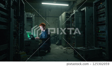 Inside a dark, cluttered server room, a technician with red hair focuses intently on a laptop screen. Surrounded by rows of old servers and equipment, the space is illuminated by the eerie glow of the 125302059