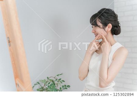 Asian woman looking at the mirror with a smile after her acne, rough skin and pimples are cured Asian woman looking at the mirror with a smile after her acne, rough skin and pimples are cured 125302894