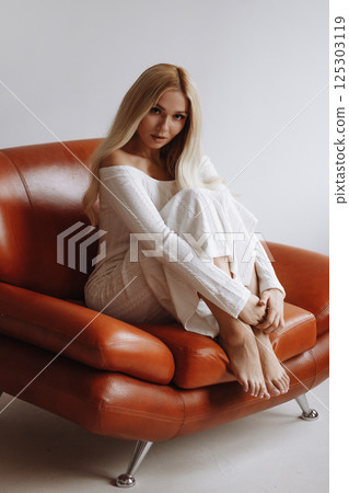 Slender blonde in a white dress sits barefoot on a leather chair against a white wall 125303119