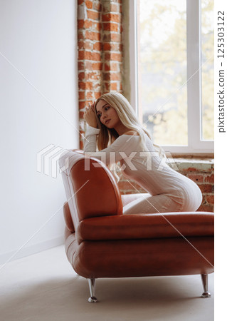 Slender blonde in a white dress sits barefoot on a leather chair against a white wall Slender blonde in a white dress sits barefoot on a leather chair against a white wall 125303122