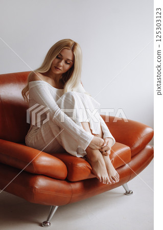 Slender blonde in a white dress sits barefoot on a leather chair against a white wall Slender blonde in a white dress sits barefoot on a leather chair against a white wall 125303123