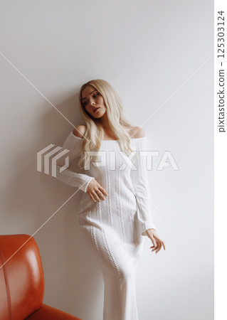 Elegant blonde in a white knitted dress stands near a white wall Elegant blonde in a white knitted dress stands near a white wall 125303124