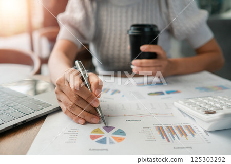 Close-up of a person reviewing financial charts with a laptop, calculator, and coffee in a modern office environment. Close-up of a person reviewing financial charts with a laptop, calculator, and coffee in a modern office environment. 125303292