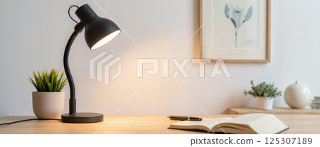 A desk lamp illuminates a wooden office desk with an open book, a potted plant, a pen, and framed art on the wall. Minimalist decor with neutral colors and natural elements 125307189