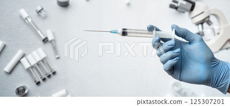 A hand in a blue glove holds a syringe, surrounded by scattered laboratory equipment, including vials and a microscope, in a clinical setting. No text is present in the image 125307201
