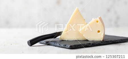 Two wedges of pale cheese with small holes rest on a dark slate board, accompanied by a sharp cheese knife with a black handle, against a light background Two wedges of pale cheese with small holes rest on a dark slate board, accompanied by a sharp cheese knife with a black handle, against a light background 125307211