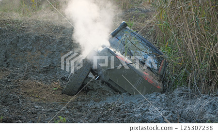 A close-up of an off-road vehicle prepared for competition, stuck in a muddy pit. The SUV is tilted at an angle, its wheels are buried in mud, and steam is rising from the engine A close-up of an off-road vehicle prepared for competition, stuck in a muddy pit. The SUV is tilted at an angle, its wheels are buried in mud, and steam is rising from the engine 125307238
