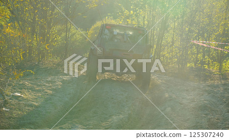 A muddy SUV drives through the sand with dense woods kicking up dust. Sunlight streams through the trees, casting spectacular shadows. Rear view. Extreme off-road racing 125307240