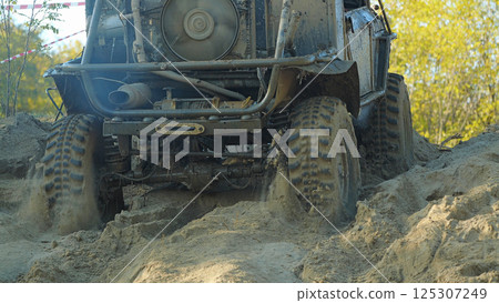 A muddy off-road vehicle navigates a challenging sandy terrain. The vehicle's tires are deeply embedded in the sand, and the suspension is flexed as it climbs the slope A muddy off-road vehicle navigates a challenging sandy terrain. The vehicle's tires are deeply embedded in the sand, and the suspension is flexed as it climbs the slope 125307249