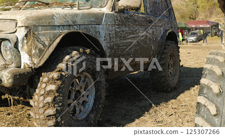 A close-up of a muddy off-road vehicle with large, knobby tires. The vehicle is covered in mud. Other vehicles and spectators are visible in the background A close-up of a muddy off-road vehicle with large, knobby tires. The vehicle is covered in mud. Other vehicles and spectators are visible in the background 125307266