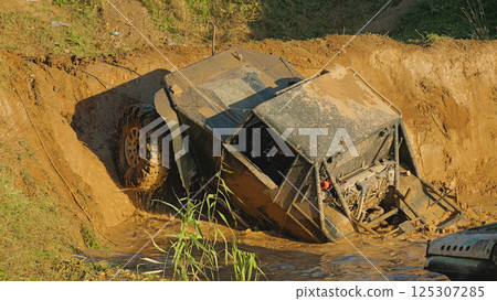 A muddy off-road vehicle designed for extreme off-roading partially peeks out of a deep puddle. The rear part of the vehicle is in water. There is a steep dirt slope in front of it A muddy off-road vehicle designed for extreme off-roading partially peeks out of a deep puddle. The rear part of the vehicle is in water. There is a steep dirt slope in front of it 125307285