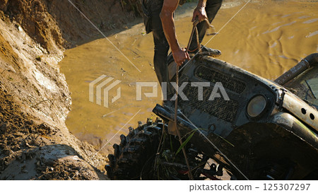 A male crew navigator stands on the hood of an SUV stuck in a muddy ditch and pulls the winch rope. Extreme racing A male crew navigator stands on the hood of an SUV stuck in a muddy ditch and pulls the winch rope. Extreme racing 125307297