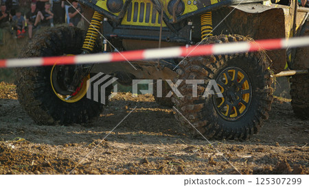 Off-road vehicle on an obstacle course. Close-up of an SUV with large mud tires, suspension and frame. The vehicle is standing near a barrier with red and white stripes Off-road vehicle on an obstacle course. Close-up of an SUV with large mud tires, suspension and frame. The vehicle is standing near a barrier with red and white stripes 125307299