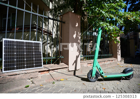 Mint-green electric scooter parked on stone pavement next to solar panel leaning against iron fence. Solar panel connected to scooter, emphasizing sustainable method of charging in urban setting. Mint-green electric scooter parked on stone pavement next to solar panel leaning against iron fence. Solar panel connected to scooter, emphasizing sustainable method of charging in urban setting. 125308313