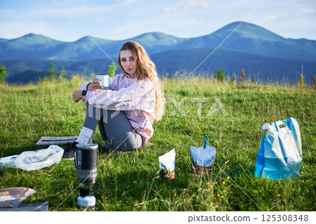 Woman tourist sits on grassy hillside, holding cup. Traveler with long blonde hair. Nearby portable camping stove, food pouches, blue bag. Scenic mountain backdrop adds to serene outdoor atmosphere. 125308348