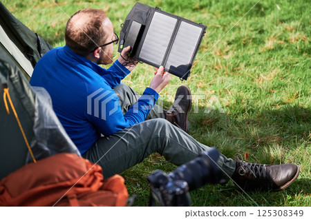 Man sits beside tourist tent, holding portable solar panel charger to capture sunlight for charging power bank. Renewable energy in outdoor activities, blending technology with natural environment. 125308349