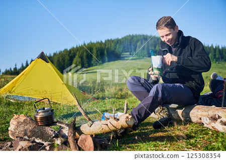 Man tourist eating from food pouch with green spoon, sits on log near campfire. Yellow tent on green hillside, surrounded by lush trees. Peacefulness of camping in beautiful natural environment. 125308354