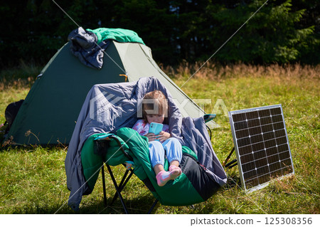 Young child plays with smartphone while charging with photovoltaic solar panel near tourist tent in summer. Integration of renewable energy in outdoor camping activities. Young child plays with smartphone while charging with photovoltaic solar panel near tourist tent in summer. Integration of renewable energy in outdoor camping activities. 125308356