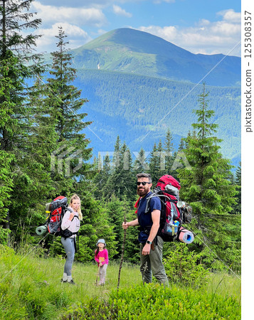 Family of three, two adults carrying large backpacks and child in pink jacket and hat, pauses on grassy hillside. Stunning mountain landscape with lush green trees and clear blue sky on background. 125308357