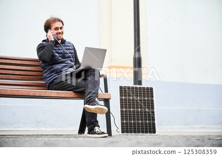 Man sits on wooden bench, working on laptop and smartphone, both connected to solar panel. Urban setting and casual attire highlight modern, sustainable, mobile workspace. Man sits on wooden bench, working on laptop and smartphone, both connected to solar panel. Urban setting and casual attire highlight modern, sustainable, mobile workspace. 125308359
