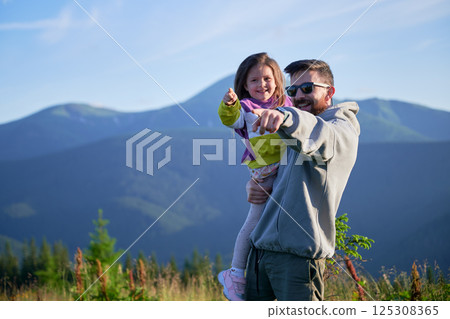 Father and his young daughter, both smiling, point towards horizon as they enjoy scenic mountain view. Father holds his daughter in his arms. Lush greenery and distant mountains under clear blue sky. 125308365