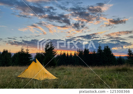Yellow tourist tent set up in grassy field at sunset, surrounded by trees in evening. Sky painted with vibrant hues of orange, pink, and purple as sun sets, casting warm glow over serene campsite. 125308366