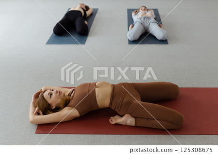 Slim young woman lying in Supta Virasana, reclining Hero Pose, stretching abdominal and back muscles on mat in gym 125308675
