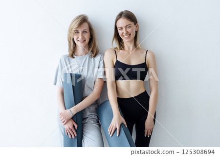 A group of young sports girls with yoga mats standing against a white wall. Girlfriends in the gym relaxing after fitness or yoga, indoors 125308694