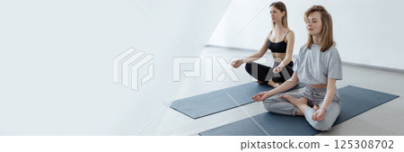 Two athletic attractive girls sit in lotus position on yoga mat indoors .Yoga, fitness and healthy lifestyle Two athletic attractive girls sit in lotus position on yoga mat indoors .Yoga, fitness and healthy lifestyle 125308702
