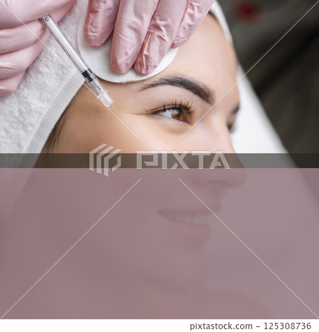 Close up of hands of young cosmetologist injecting botox in female face. She is standing and smiling. The woman is closed her eyes with relaxation 125308736