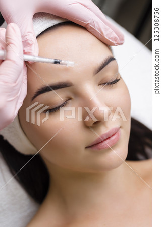Close up of hands of young cosmetologist injecting botox in female face. She is standing and smiling. The woman is closed her eyes with relaxation Close up of hands of young cosmetologist injecting botox in female face. She is standing and smiling. The woman is closed her eyes with relaxation 125308756