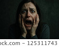 Caught in a moment of intense fear, a young woman sits in a dimly lit room with her hands on her face, mouth open in a silent scream. Her wide eyes reflect panic and despair, surrounded by shadows 125311743