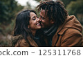 Two individuals are embracing each other in a picturesque wooded area, surrounded by trees displaying vibrant fall foliage. They are wearing warm, stylish coats and appear to be enjoying each other's 125311768