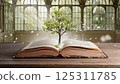An open book rests on a wooden table in a tranquil library, with soft light streaming through large, arched windows. A small green tree emerges from the center of the pages, symbolizing growth and 125311785