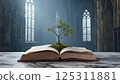 Inside a grand gothic cathedral, sunlight filters through intricate stained glass windows, casting a serene light on an open ancient book. At the center of its pages, a small green tree emerges 125311881