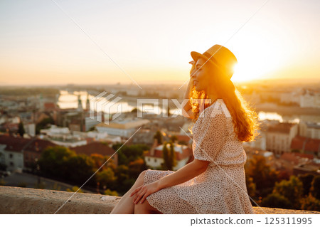 Smiling female tourist in hat admires landscape of city at dawn. Euro-trip. Active lifestyle concept 125311995