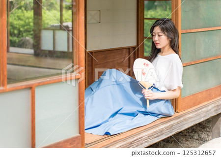 一位年輕女子坐在古民居的門廊上,用扇子搧風降溫 一位年輕女子坐在古民居的門廊上,用扇子搧風降溫 125312657