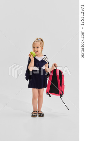 Smiling girl in navy pleated uniform eating green apple while holding backpack in one hand against white background. Smiling girl in navy pleated uniform eating green apple while holding backpack in one hand against white background. 125312971