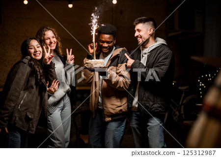 Friends celebrating with a cake and sparkler at a cozy indoor gathering during an evening event Friends celebrating with a cake and sparkler at a cozy indoor gathering during an evening event 125312987