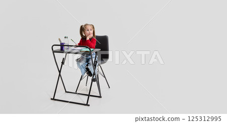 Girl in red sweater sitting at desk with hand on chin, looking away thoughtfully, surrounded by notebooks and pencils. 125312995