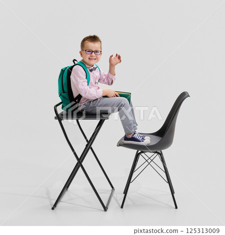 Boy in glasses wearing pink shirt and bow tie holding book and smiling while sitting on desk with backpack. Concept of confident student 125313009