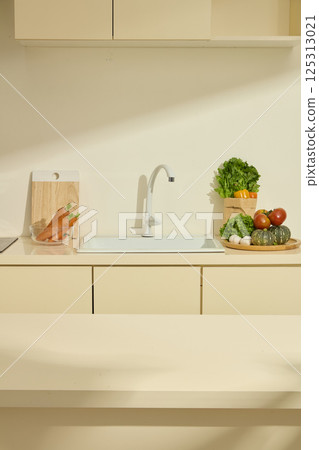 Beige empty table for products or dinner. Sink on the back counter, flanked by glass bowls of carrots. Round wooden plate with organic vegetables. Bunch of greens standing in a recycled paper bag. 125313021
