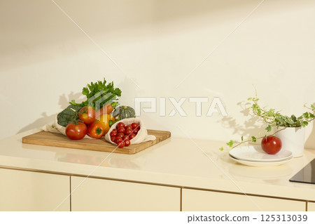 Large wooden cutting board on the kitchen counter with vegetables stacked on top. White ceramic plate with tomato on top. Green vines in a ceramic pot, decoration in a modern kitchen. Copy space. 125313039