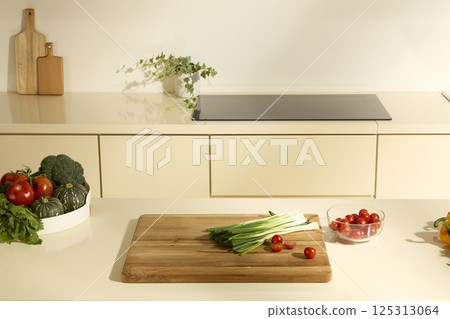 Green onions and tomatoes cut in half are placed on a wooden cutting board in the kitchen. Pumpkin, cauliflower and tomatoes are placed on a white plate. Potted plants are placed next to a cooker. Green onions and tomatoes cut in half are placed on a wooden cutting board in the kitchen. Pumpkin, cauliflower and tomatoes are placed on a white plate. Potted plants are placed next to a cooker. 125313064