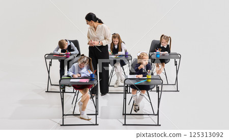 Focused children writing with colorful pens at individual desks as teacher supervises classroom activity. Concept of school task Focused children writing with colorful pens at individual desks as teacher supervises classroom activity. Concept of school task 125313092