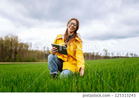 Young farmer woman in a green field, intently using a tablet. She wears a yellow jacket and glasses 125313243