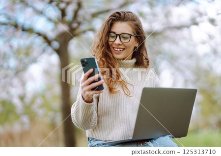 Cheerful woman on phone takes selfie while sitting at table with laptop in spring park. Lifestyle. 125313710