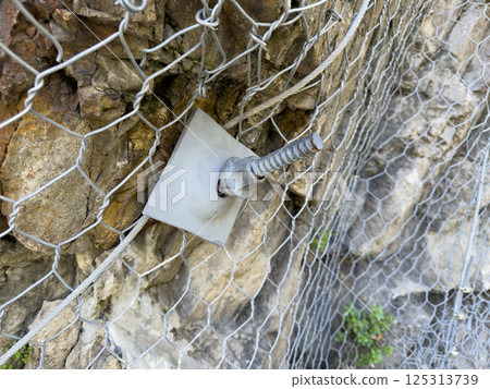 Close up of rockfall protection mesh secured with steel anchor bolt on cliffside. Industrial safety net system for landslide prevention and slope reinforcement in mountainous terrain 125313739