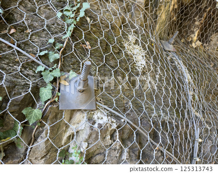 Protective metal mesh with anchored bolt plate covers steep cliff rocks. Anti rockfall safety system used in mountainous regions for slope stabilization and infrastructure defense 125313743
